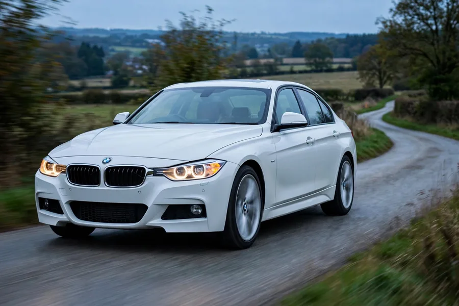White F30-generation BMW 3 Series sedan driving on a winding country road at dusk
