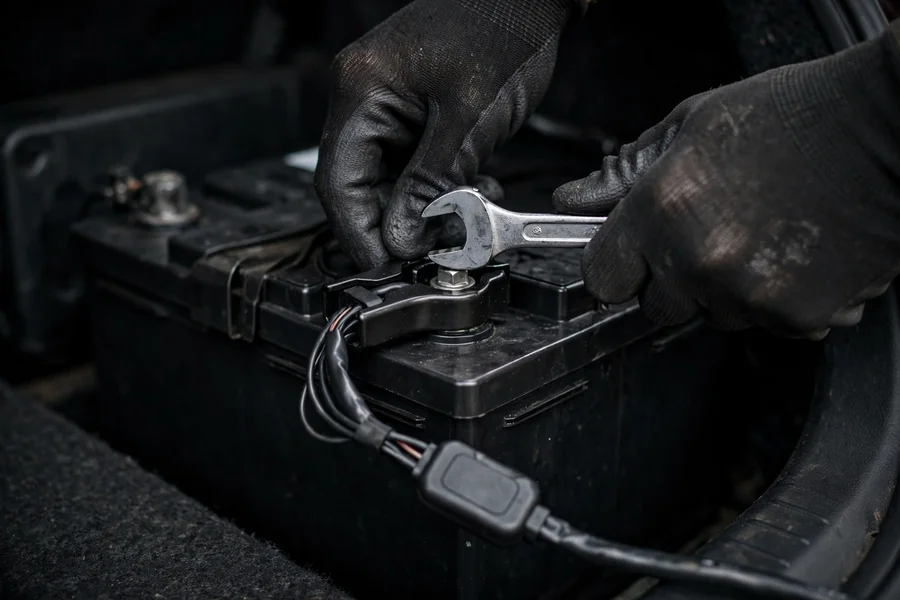 Gloved hands loosening the negative terminal clamp on a BMW 3 Series trunk battery with a wrench