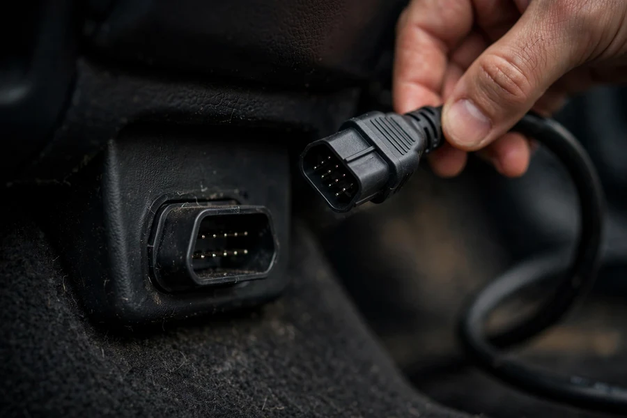 Handheld BMW-specific diagnostic scanner with cable connected to the OBD2 port beneath a car's dashboard