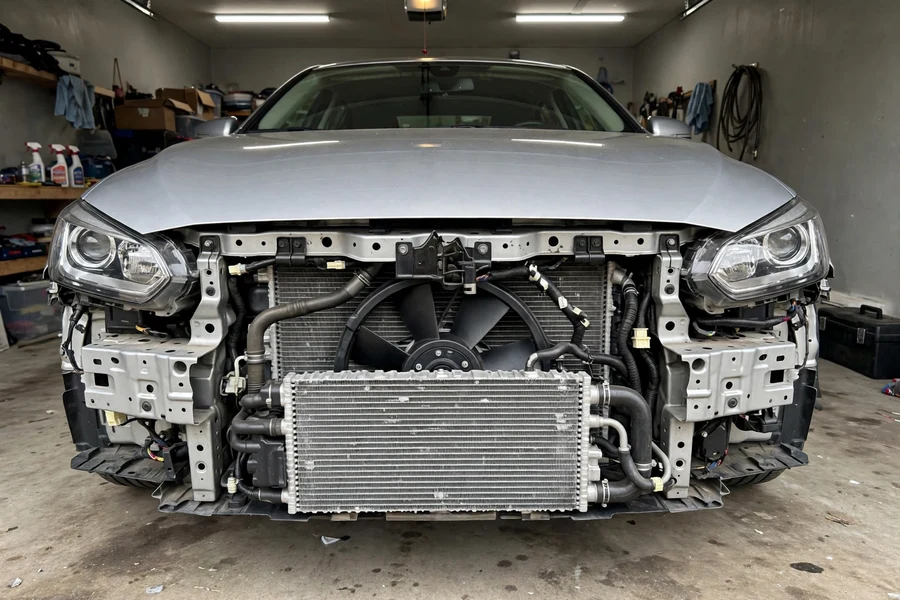 Front of a sedan with the bumper cover removed, exposing the radiator, condenser, and cooling fan during a radiator replacement